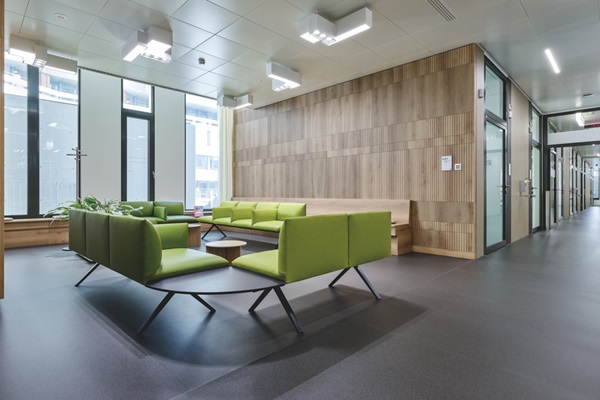 Wartebereich im Spital mit Holzwänden und -bänken, dunklem Boden und grünen Sitzpolstern